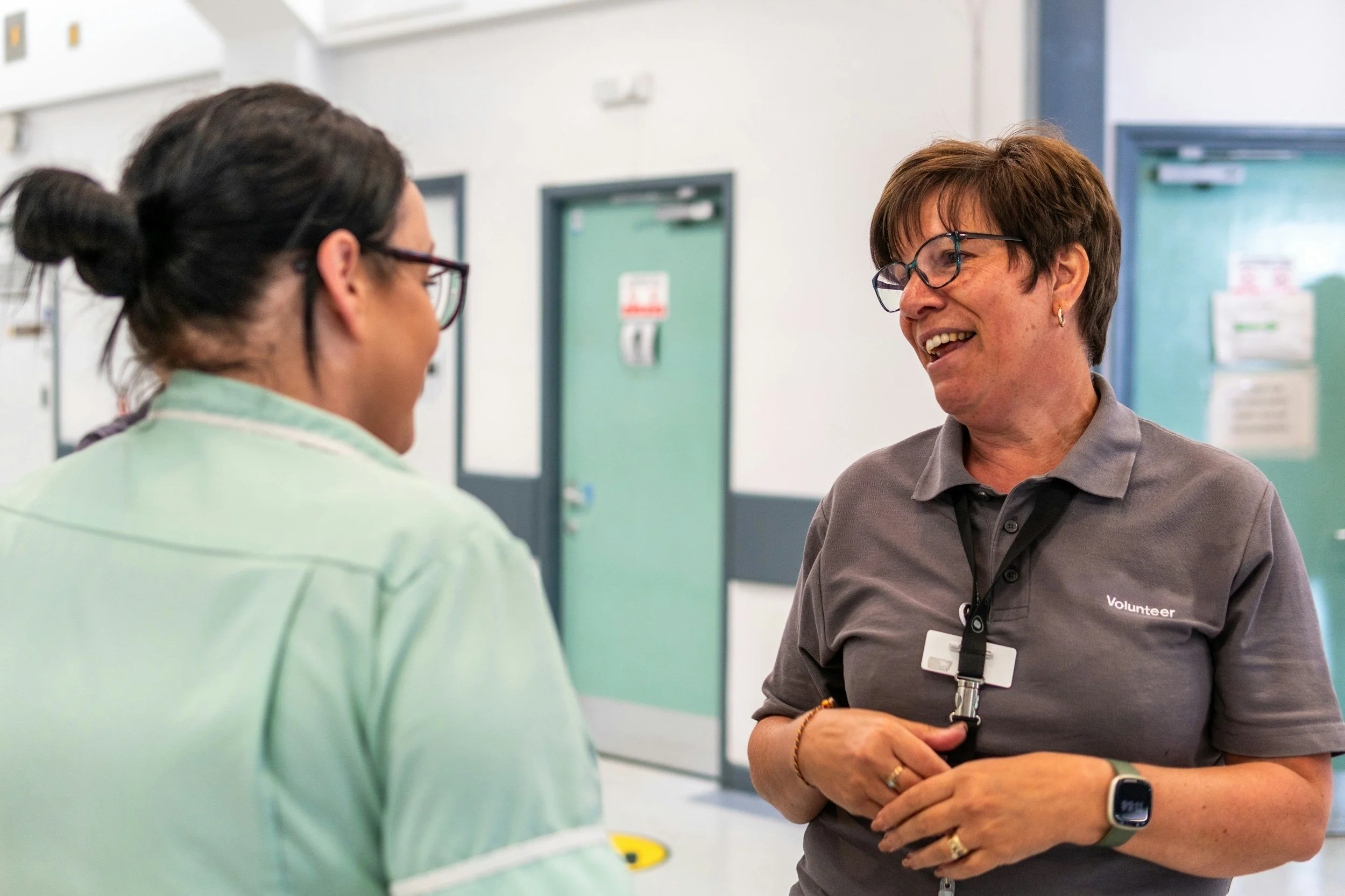 Two women, one wearing a volunteer shirt with a name badge and glasses, are smiling and conversing in an indoor setting with green doors and informational posters in the background.