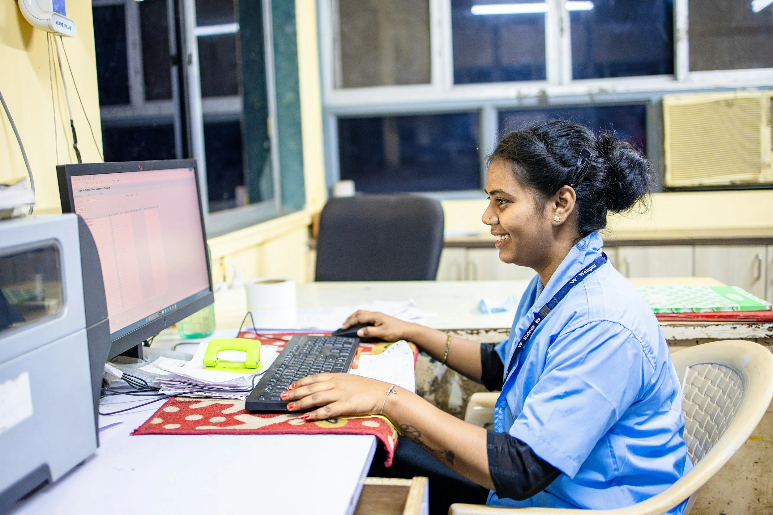 A woman with dark hair in a bun, smiling and working at a desk with a computer. She is wearing a blue shirt, a black bracelet, earrings, and a lanyard with an office or company's name. The desk has papers, a keyboard, a monitor, a yellow phone, and a polka dot cloth. The background shows a window with black curtains and a yellow air conditioner.