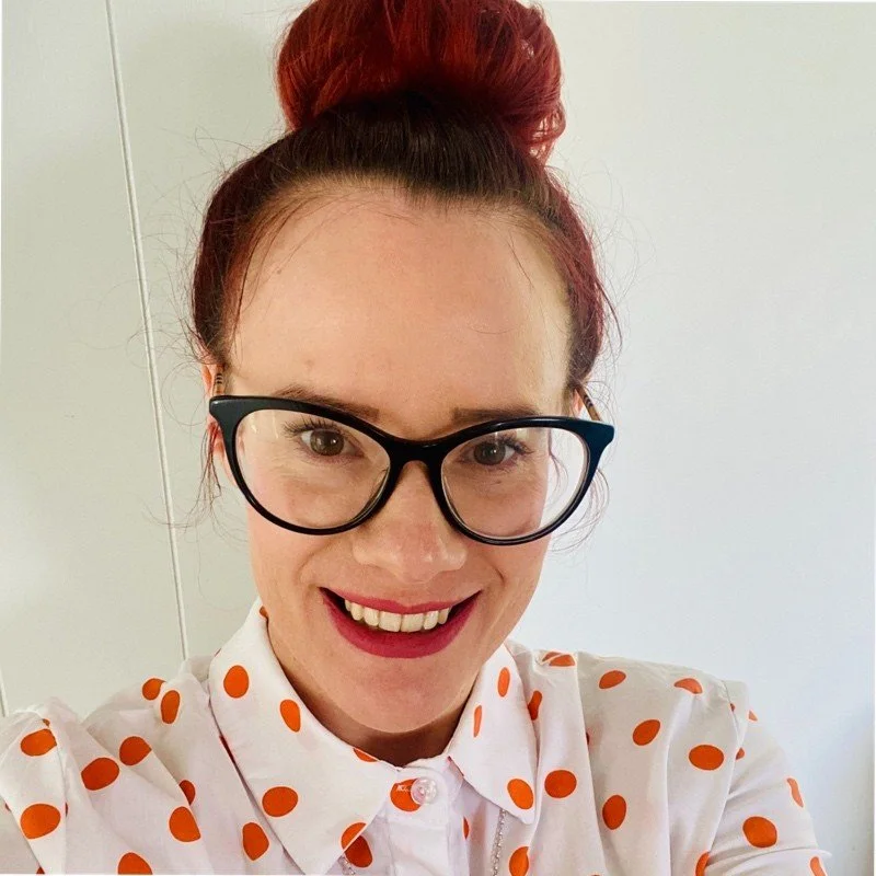 A woman with red hair tied in a bun, wearing glasses, a white shirt with orange polka dots, and smiling.