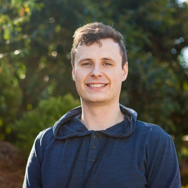 A young man with short brown hair and a friendly smile, outdoors with greenery in the background, wearing a navy blue hoodie.