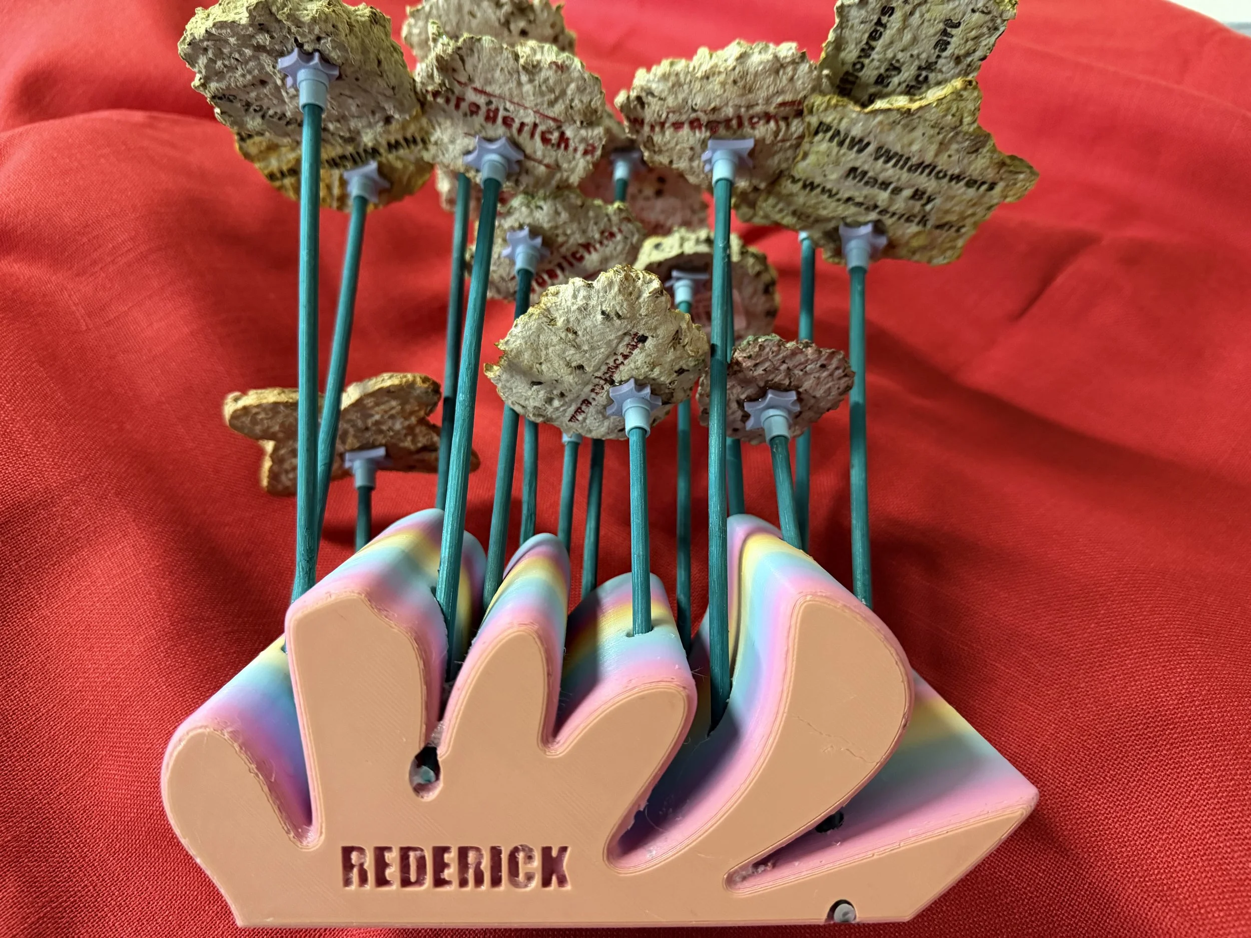 An assortment of decorative pins with colorful butterfly-shaped tops and floral cupcake toppers attached to metal sticks, displayed in a pink holder labeled 'REDDERICK' against a red fabric background.
