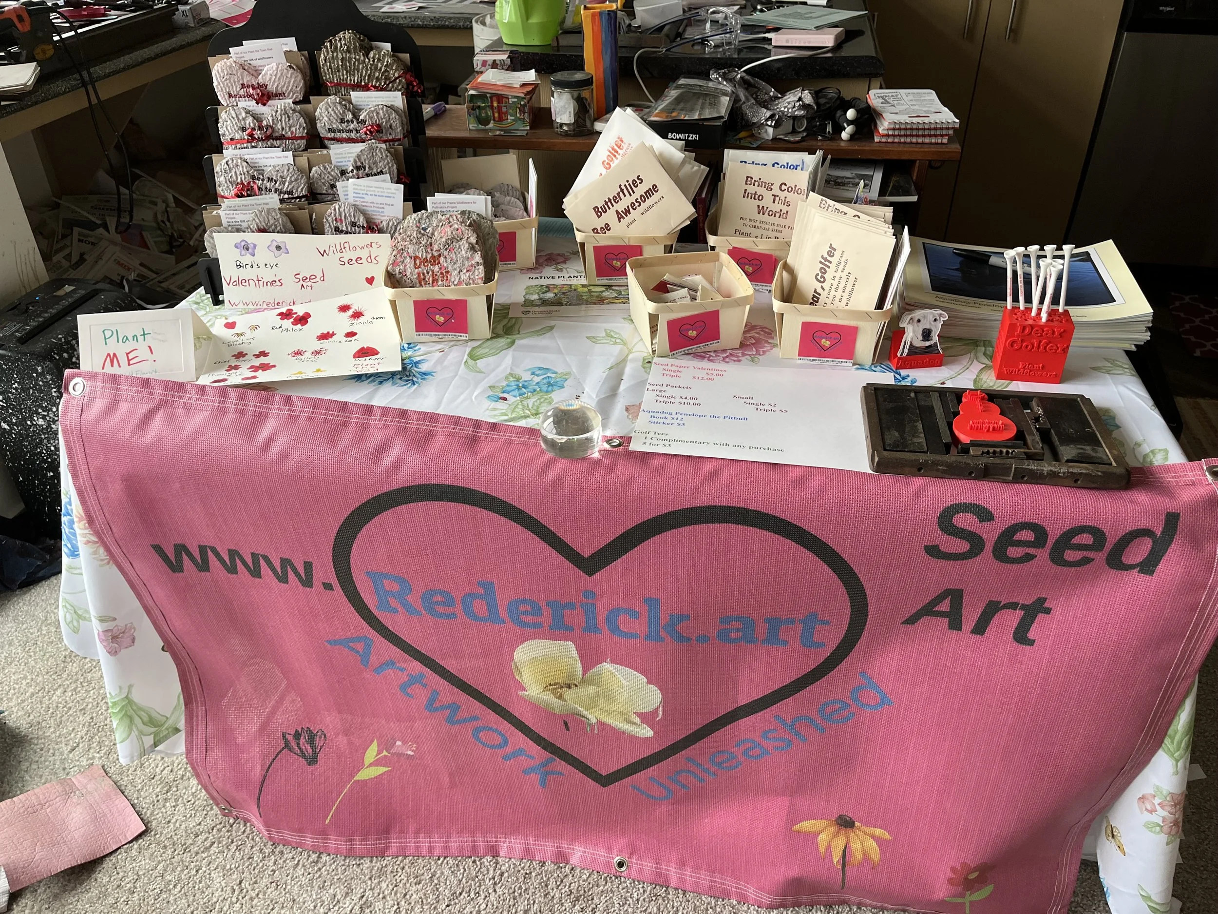 A table covered with a pink cloth displaying various seed art items, including decorated rocks and paper crafts, for sale at a Rederick.art event. The table has signs and containers labeled with different types of seeds and art, and a background of a busy workspace.