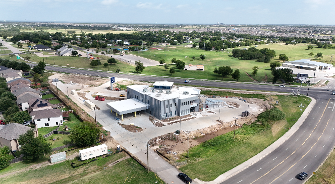 Aerial view of a new commercial building under construction near a gas station, with surrounding houses, green fields, and roads in the background.
