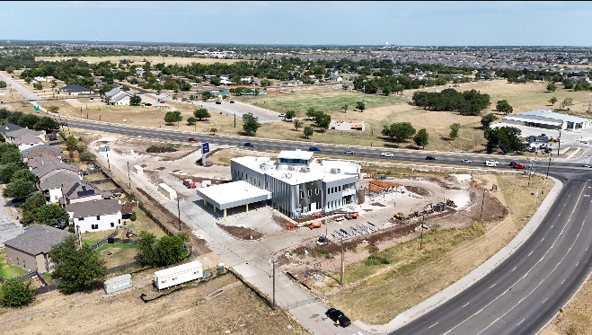 An aerial view of a construction site for a new building in a suburban area, with surrounding houses, roads, and open fields.