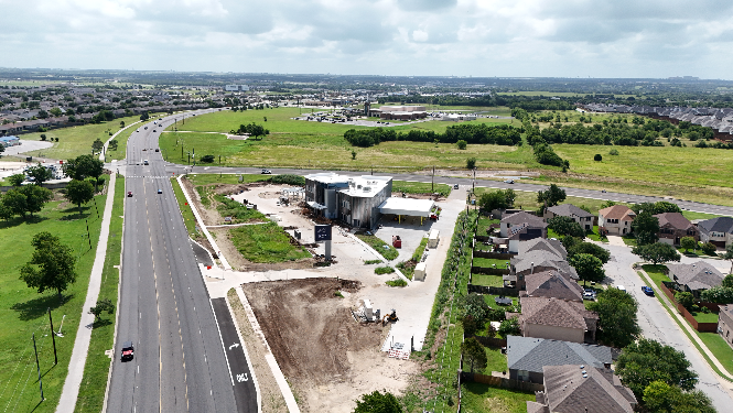 Aerial view of a suburban neighborhood with a new construction site beside a busy road, surrounded by green fields and scattered clouds.