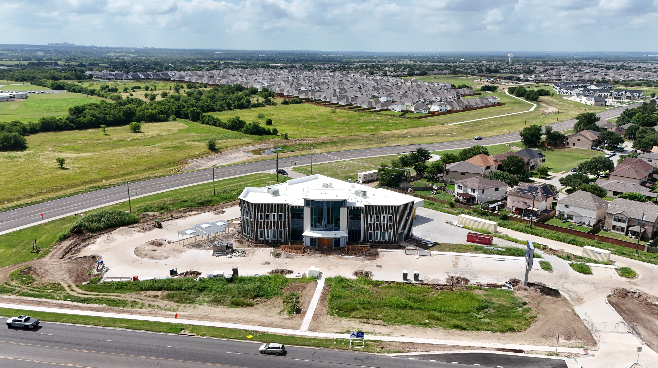 Under construction modern building with a curved facade, surrounded by dirt and grass, located near a highway with cars, residential houses, and open green fields in the background.