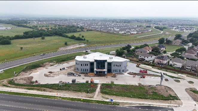 An aerial view of a construction site with a half-built circular building, surrounded by dirt, construction materials, and some vehicles. The site is near a highway, with residential houses and green fields in the background.