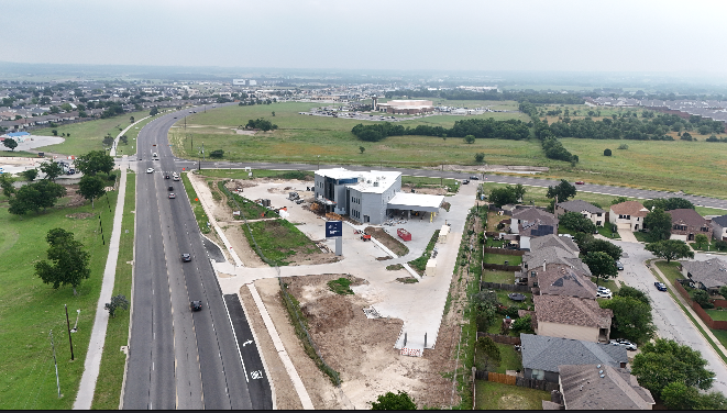 An aerial view of a developing commercial area with a modern building under construction, adjacent to a residential neighborhood and a busy highway.