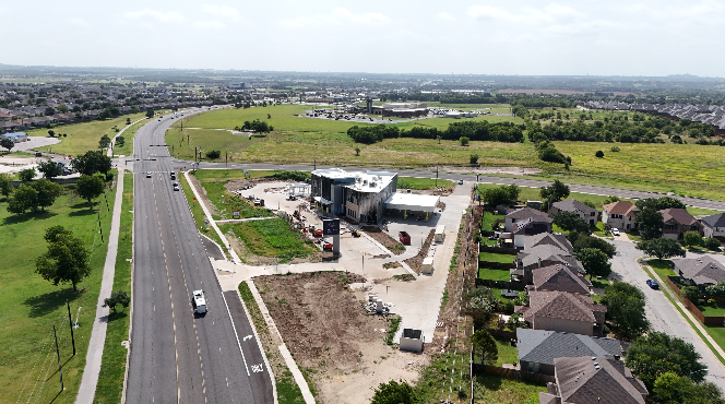 Aerial view of a developing site with a modern building, a parking lot, a residential neighborhood, and a highway in the background.