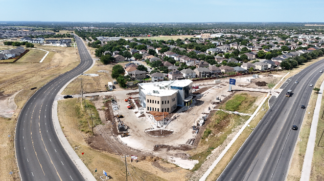 Aerial view of a construction site with a partially built white building, surrounded by dirt and construction equipment, near curved highway roads and a residential neighborhood.