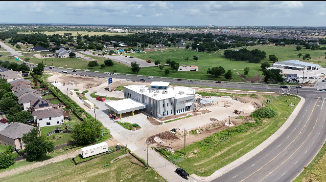 Aerial view of a new commercial building under construction in a suburban area with residential houses, green fields, and a highway in the background.