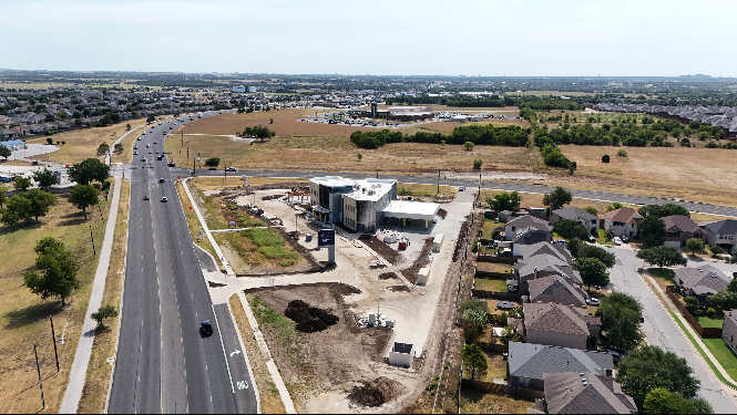 Aerial view of a suburban neighborhood with a new commercial building under construction near the main road, surrounded by houses and open fields.