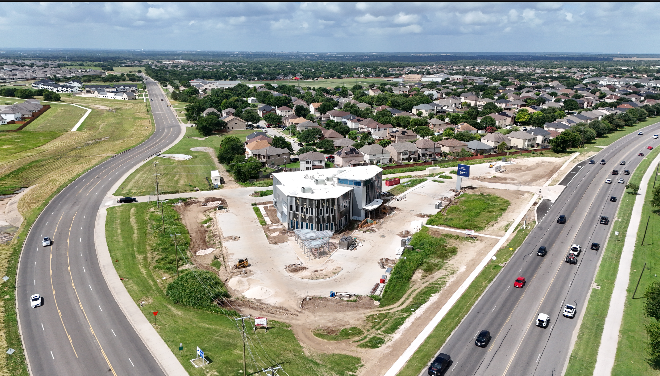 Aerial view of a construction site in a suburban area, showing a partially built commercial building surrounded by dirt, with nearby roads and residential houses in the background.