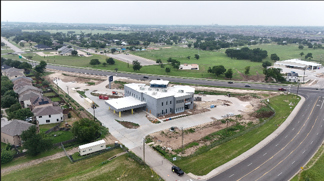 Aerial view of an unfinished modern white building under construction, surrounded by a mix of residential houses and green fields, with highway roads nearby.