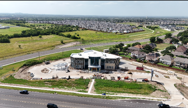 Aerial view of a construction site with a building in progress, surrounded by vehicles and dirt, with a residential neighborhood and open green fields in the background.