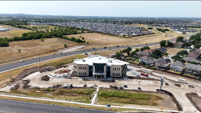 A large, modern building under construction in a suburban area, with construction equipment and materials around it and a road in front.