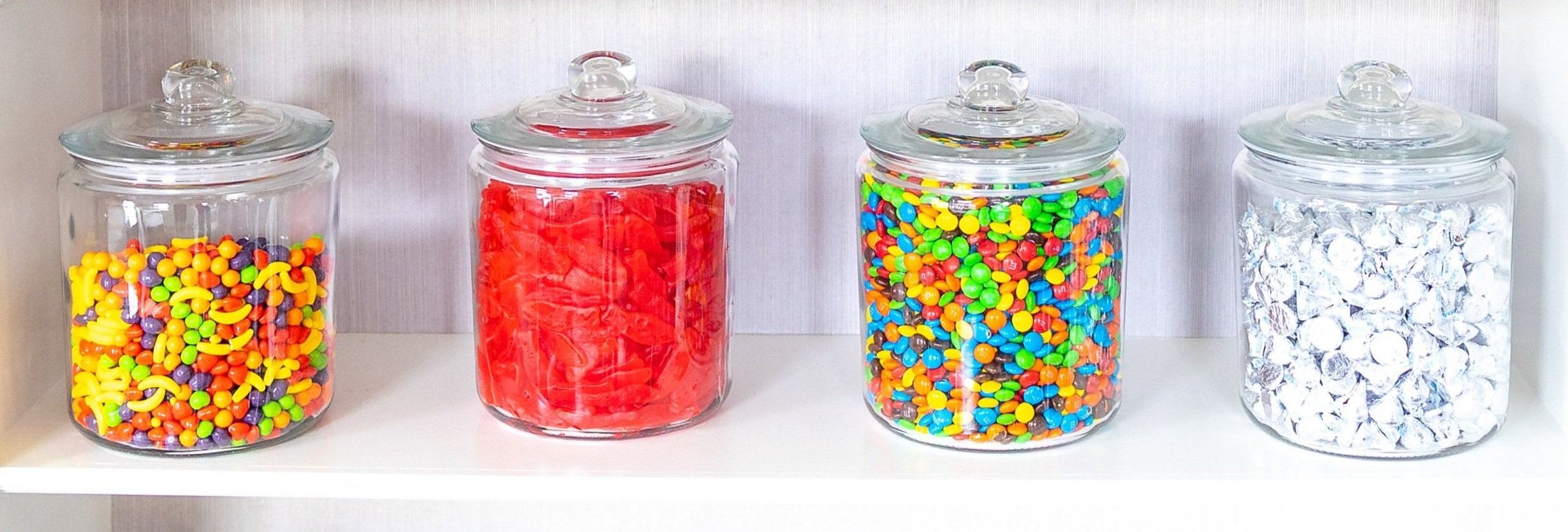 Four glass jars with lids containing colorful candies on a white shelf.