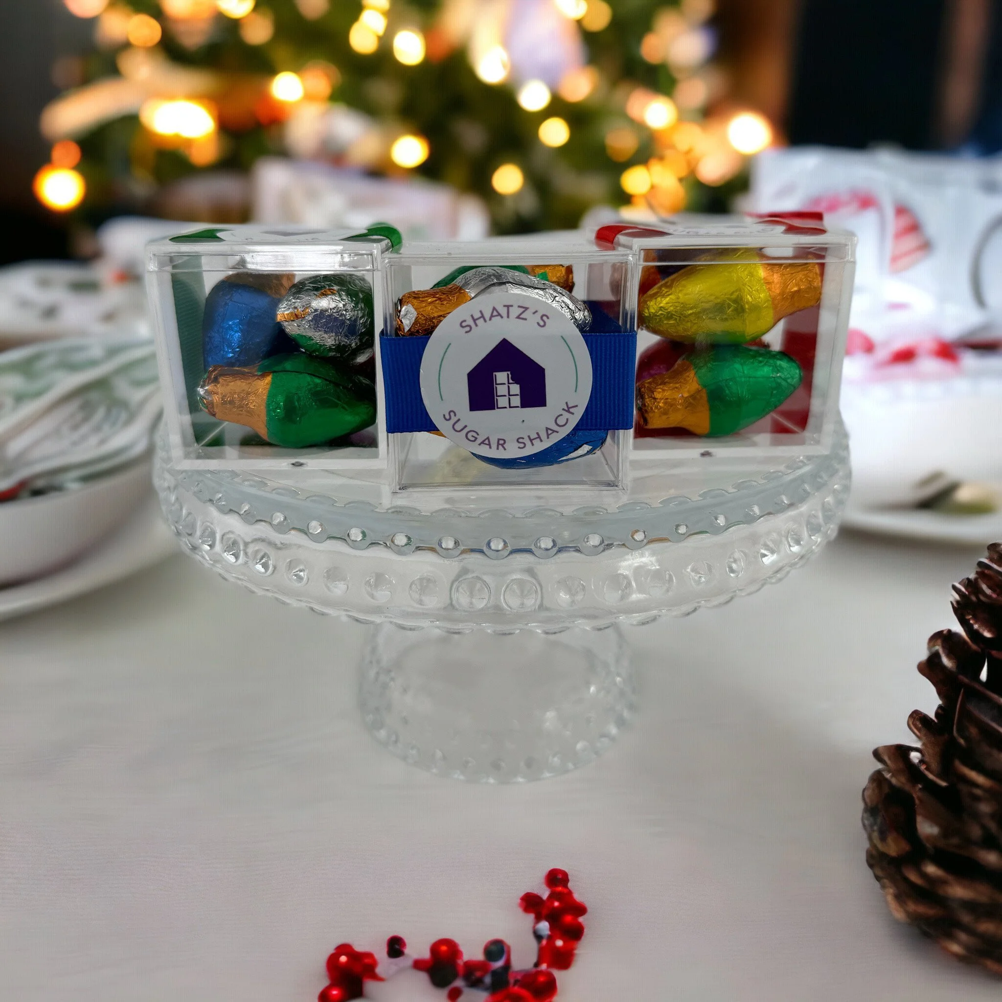 Clear glass cake stand holding three clear plastic boxes of assorted wrapped chocolates with a Christmas tree and holiday decorations in the background.