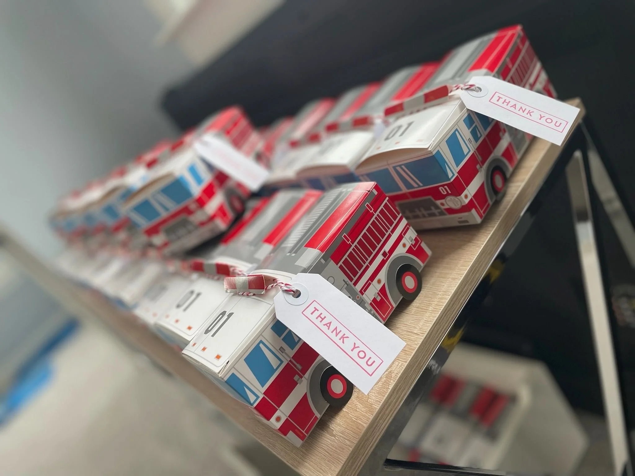 Small custom birthday party gift boxes decorated as fire trucks, lined up on a wooden shelf, each with a name tag attached.