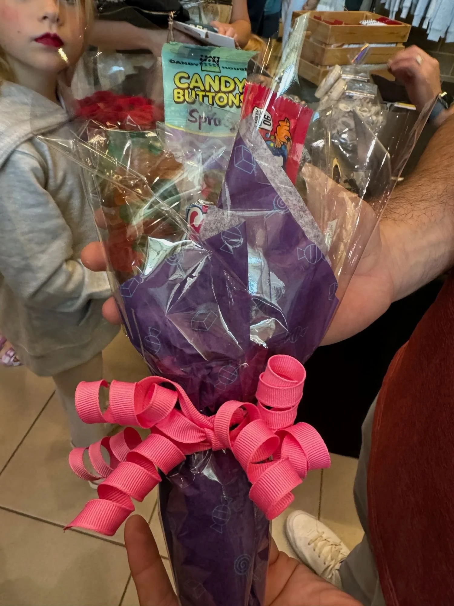Close-up of a purple-wrapped bouquet with pink curly ribbon being held by a person in a crowded indoor setting, with children and adults around.