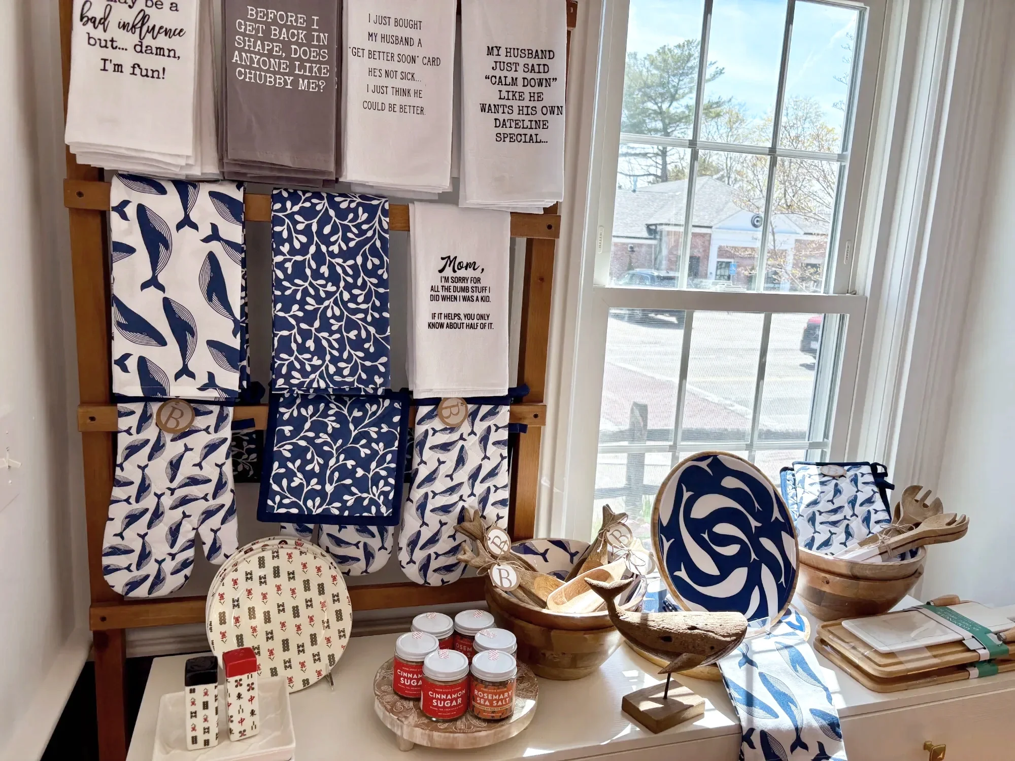 A display of kitchen towels with various patterns and phrases, some on a wooden rack and others hanging, with small decorative bowls, candles, and printed items on a table below, near a window with houses and trees visible outside.