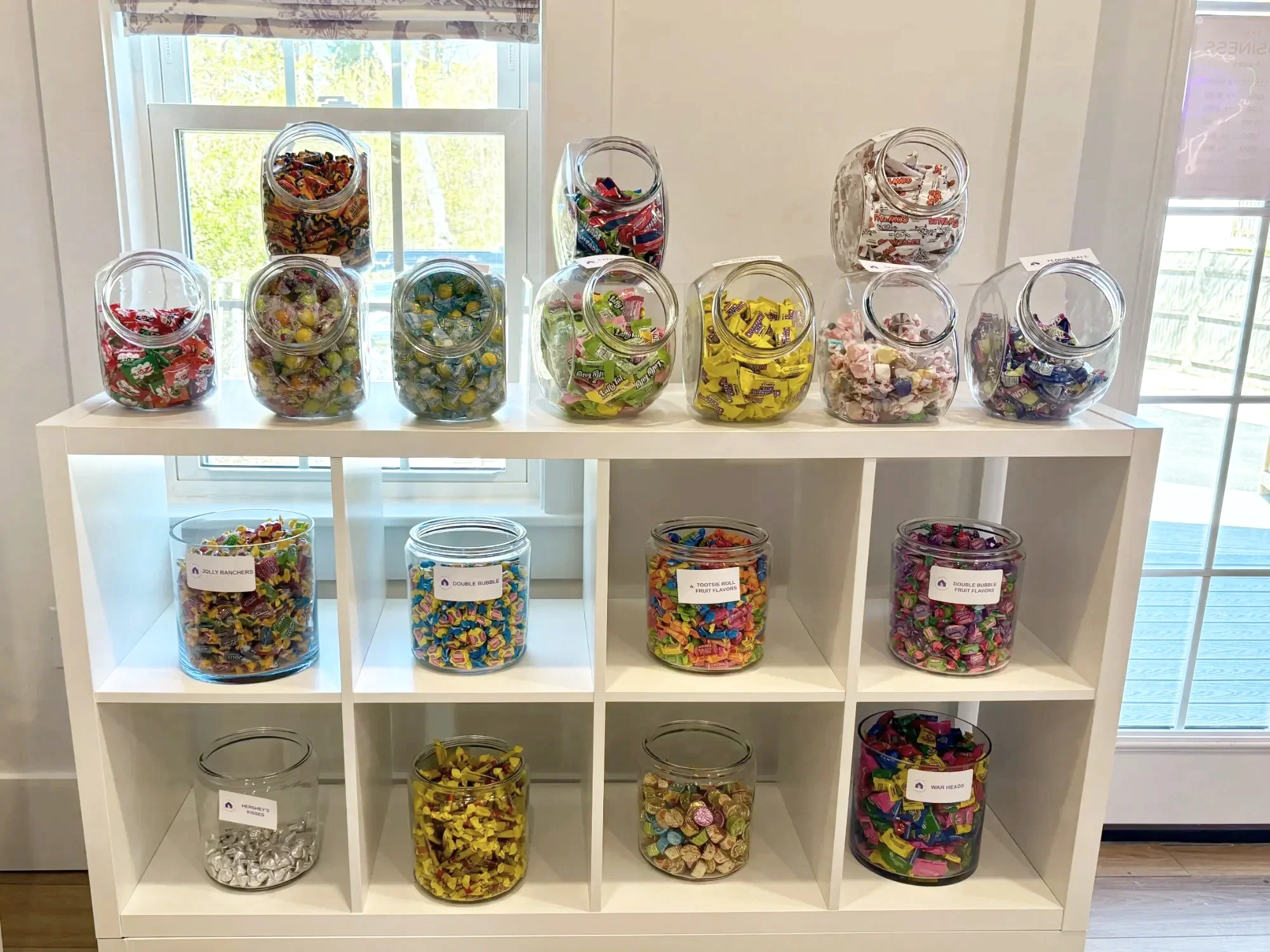 White shelf with glass jars filled with assorted candies, including wrapped chocolates and fruit-flavored chews, labeled with different names, placed in front of a window with natural light.