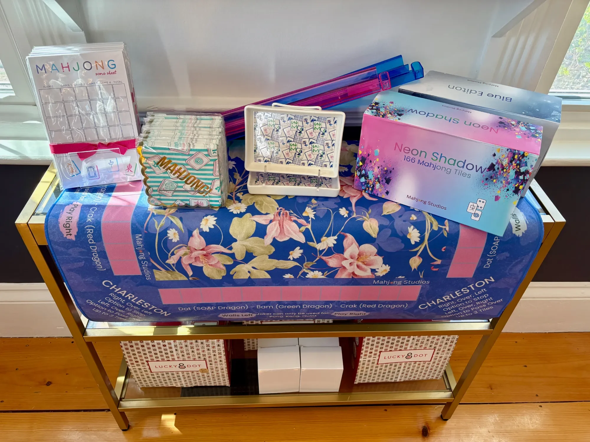 A Mahjong game set on a table with various Mahjong tiles, score sheet, and storage trays, along with a box for Mahjong tiles and a decorative tablecloth.