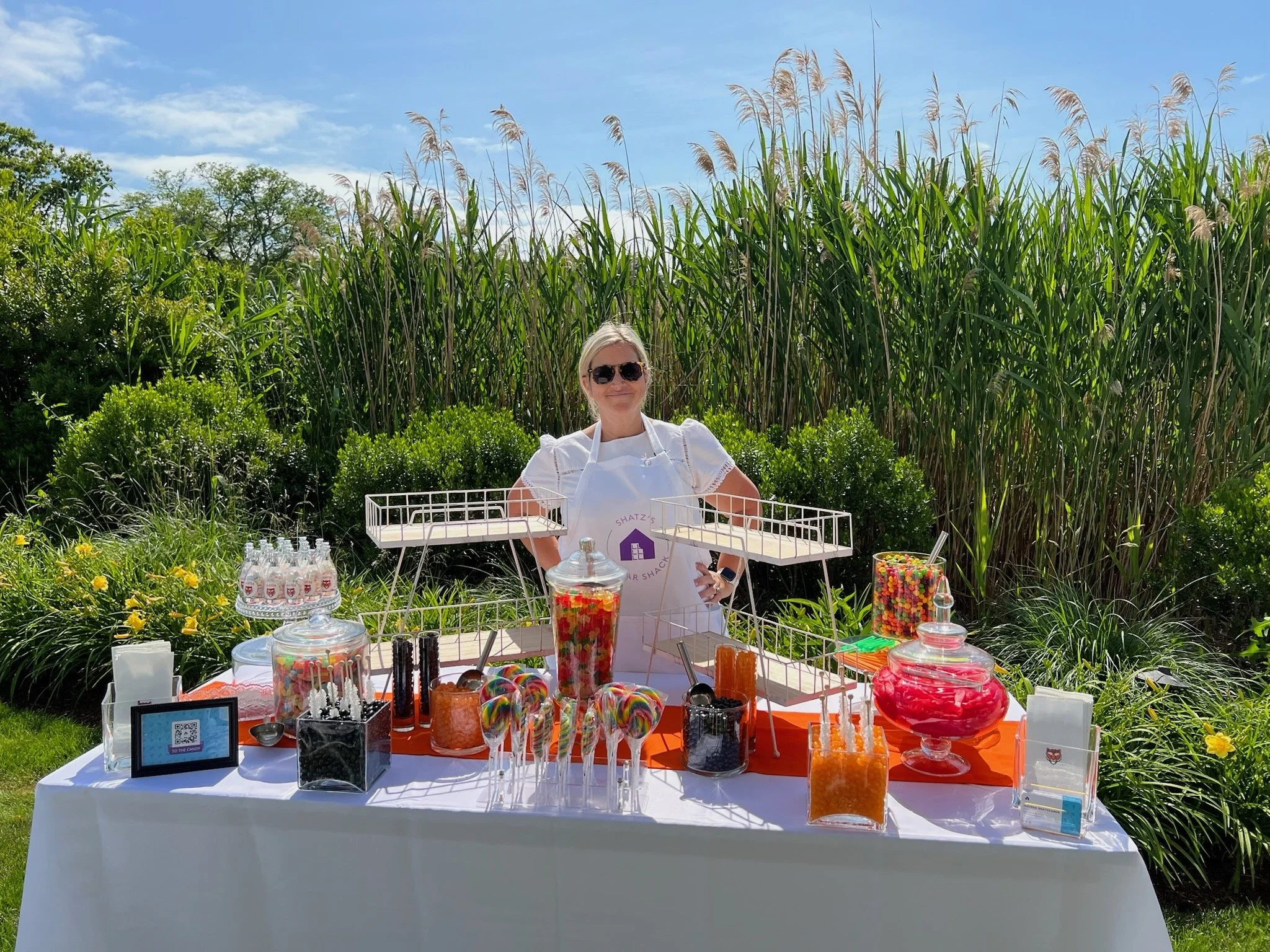Jerusha Shatzkamer, owner of Shatz's Sugar Shack, standing behind a custom candy buffet with various candies and treats, outdoors with tall grass and bushes in the background, sunny sky overhead.