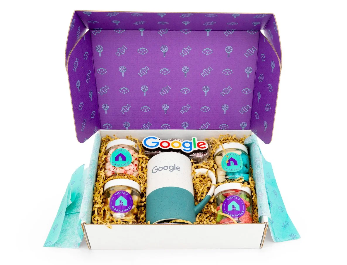 A gift box with a Google logo mug, four small jars of candy labeled "Sugar Shack," and a colorful "Google" cookie, all arranged on yellow paper shreds inside a purple box with patterned lining.