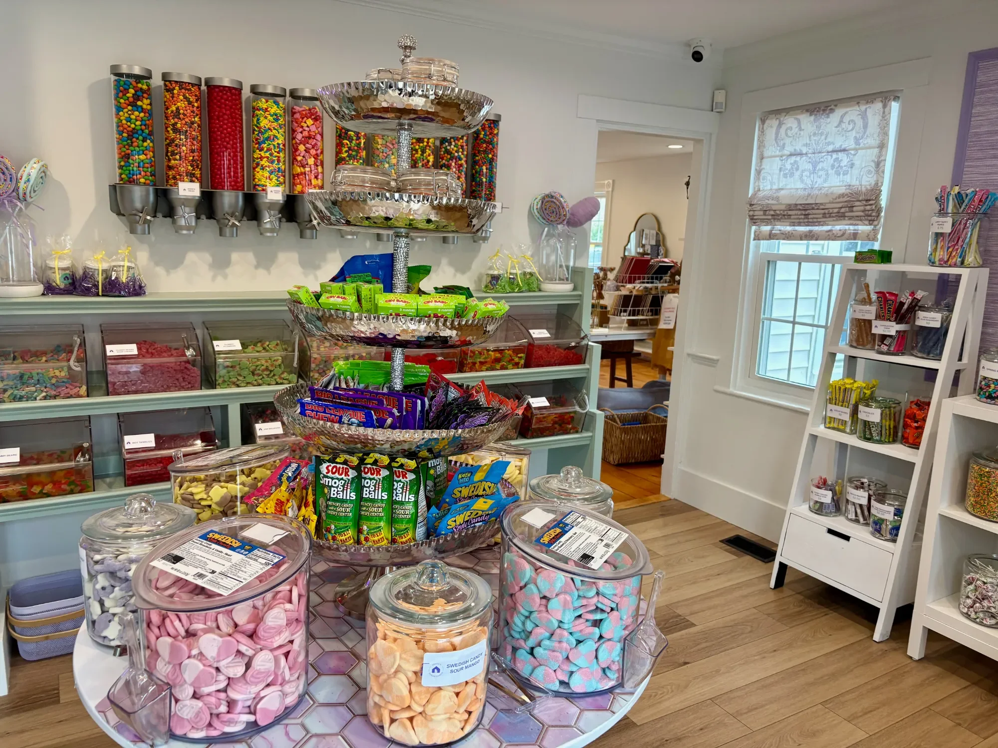 A candy shop display with various colorful candies in large glass jars and dispensers, including gummies, sour candies, marshmallows, and chocolates, with a refrigerated section in the foreground.