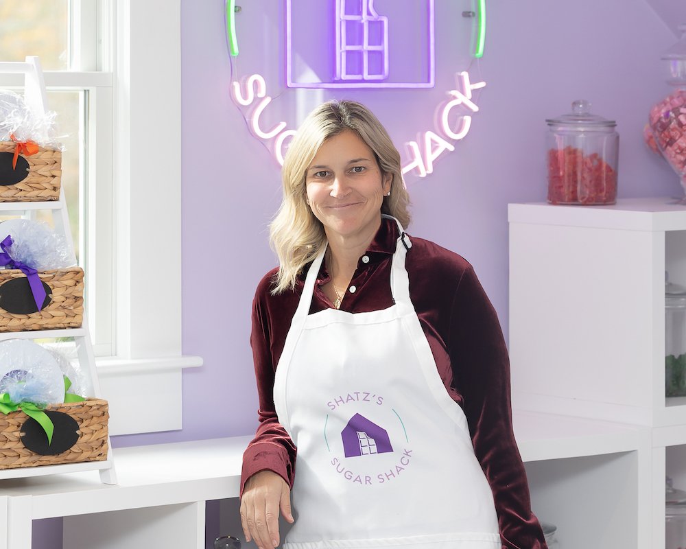 A woman wearing a white apron with a purple logo that reads "Shatz's Sugar Shack" is standing in a pastel-colored shop, smiling. Behind her are jars filled with pink and red candies, and a display shelf with baskets wrapped in clear cellophane and tied with various colored ribbons.