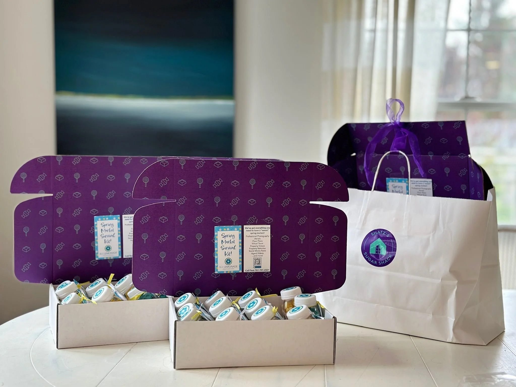 Spring Market Survival Kit with small bottles in a white box, purple boxes with open lids, and a white gift bag with purple handles and a logo, all on a white table near a window with curtains.