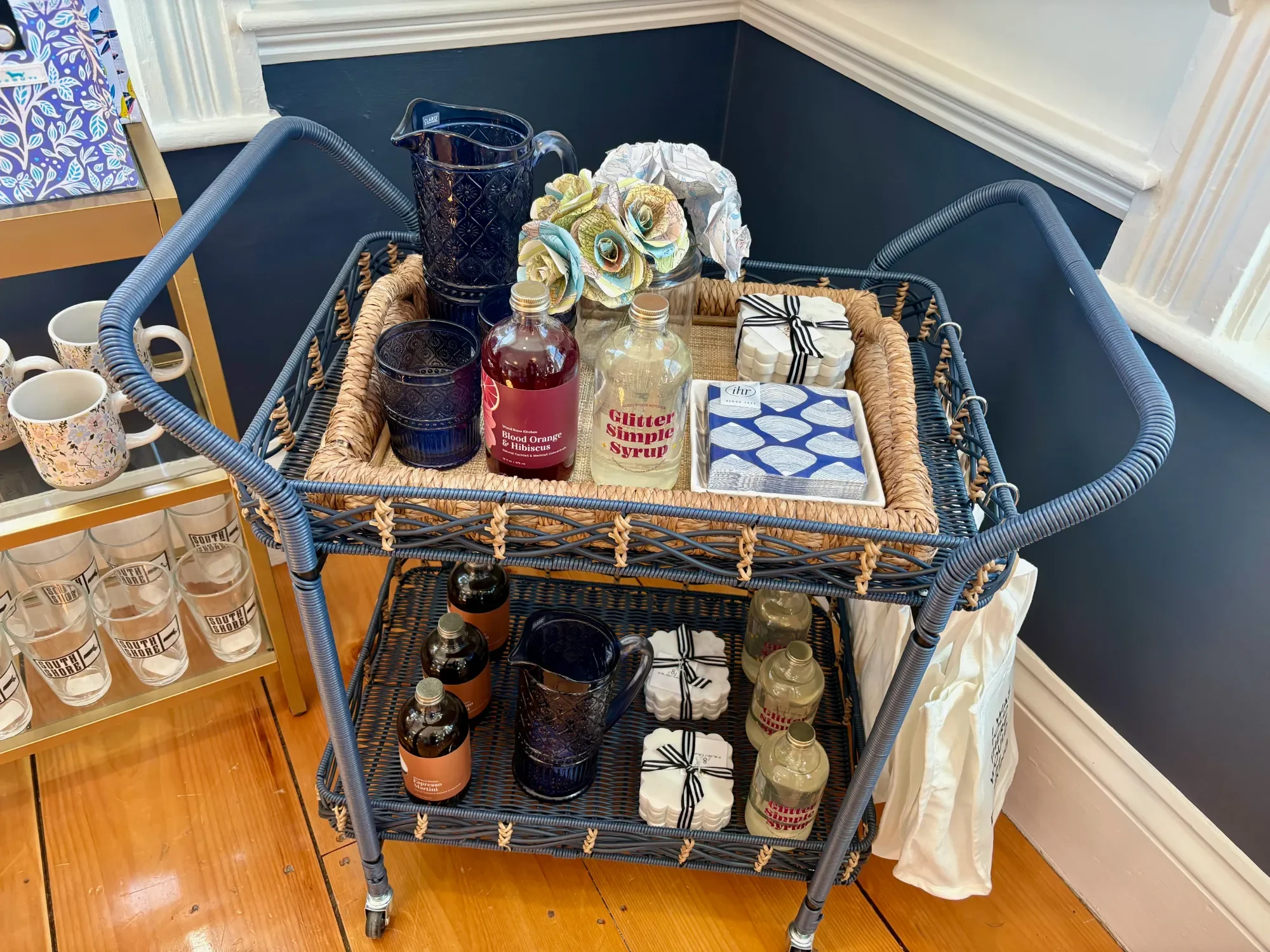 A metal and woven basket cart filled with decorative paper flowers, glassware, bottles, and packaged items, displayed in a store with wooden flooring and blue and white walls.