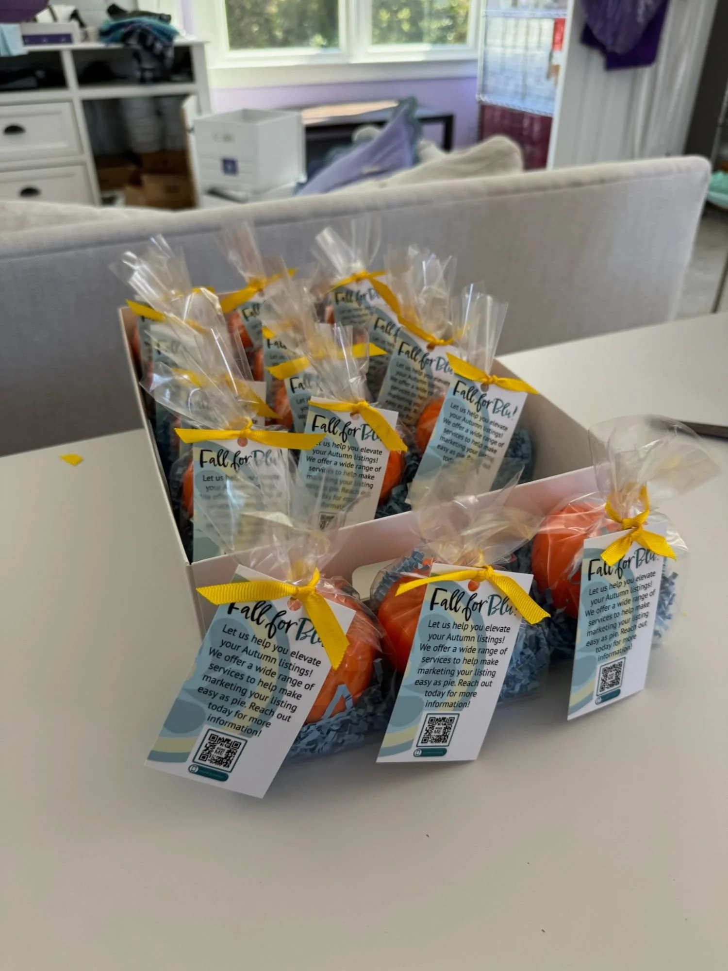 Small orange pumpkins wrapped in clear plastic with yellow ribbons, each with a tag that reads 'Fall for Blue' and a QR code, arranged in a white box on a white table.
