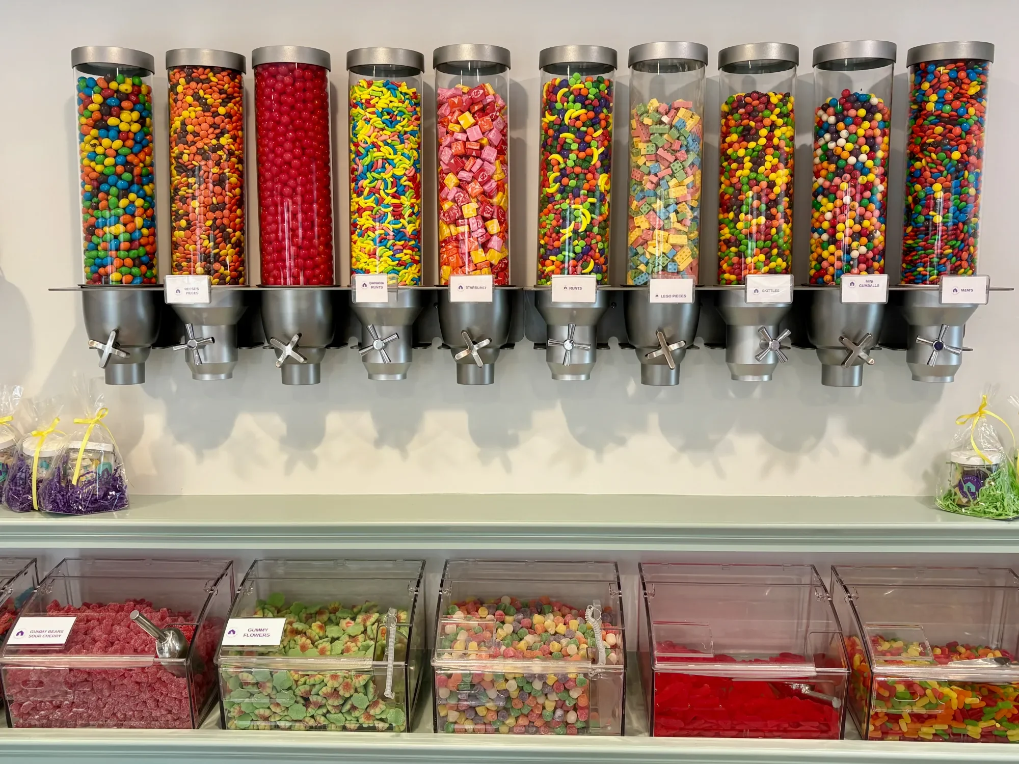 Display of various colorful candies in clear containers with metal lids and dispensing spouts, arranged in two rows.