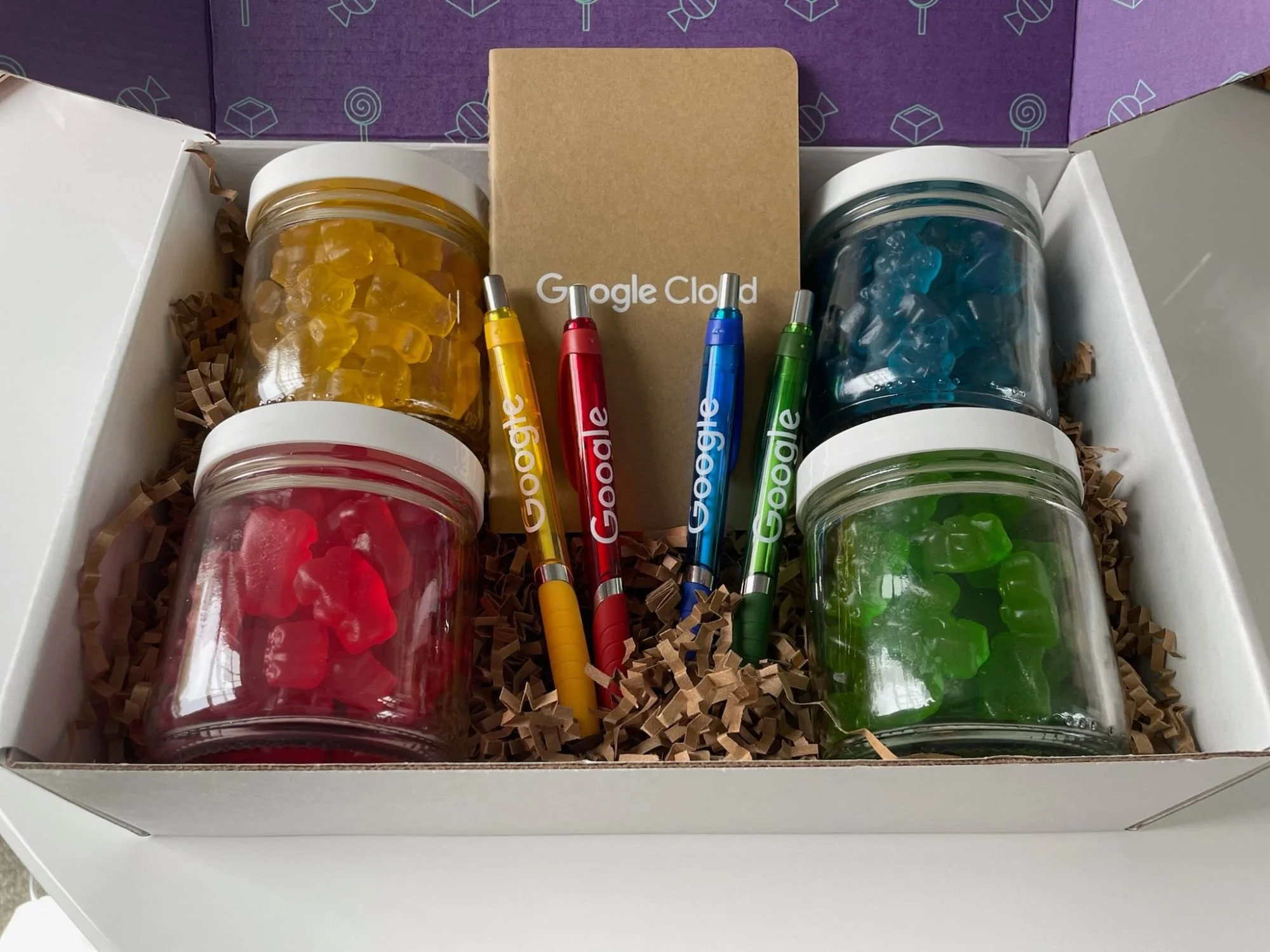 Gift box containing four jars of gummy candies in yellow, red, blue, and green colors, four Google-branded pens in yellow, red, blue, and green, and a cardboard note with 'Google Cloud' written on it, all inside a white box with brown paper filler.