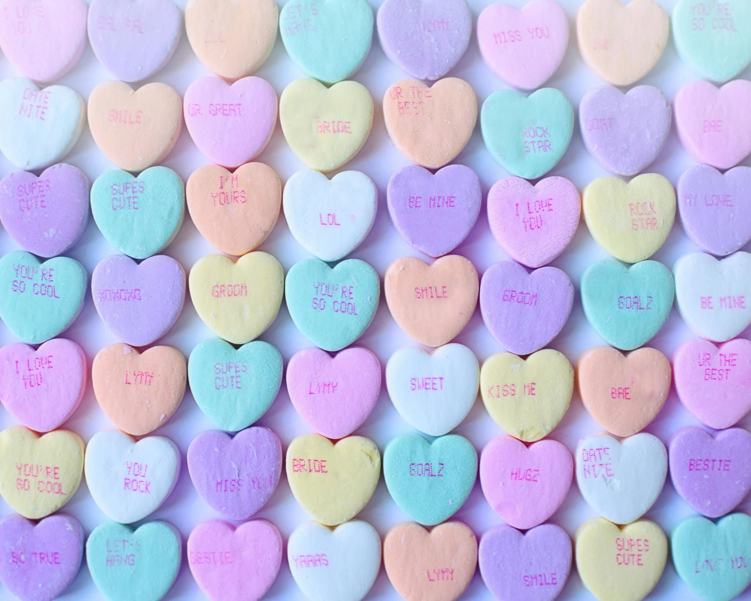 Colorful heart-shaped candies with various affectionate messages and compliments written on them in pink and purple ink, arranged in a grid pattern.
