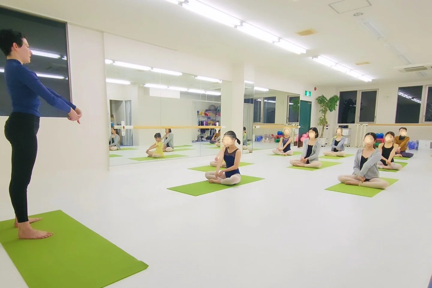 Body Tuning and conditioning class with instructor leading students seated in cross-legged pose on green mats in a dance studio with mirrors and large windows.