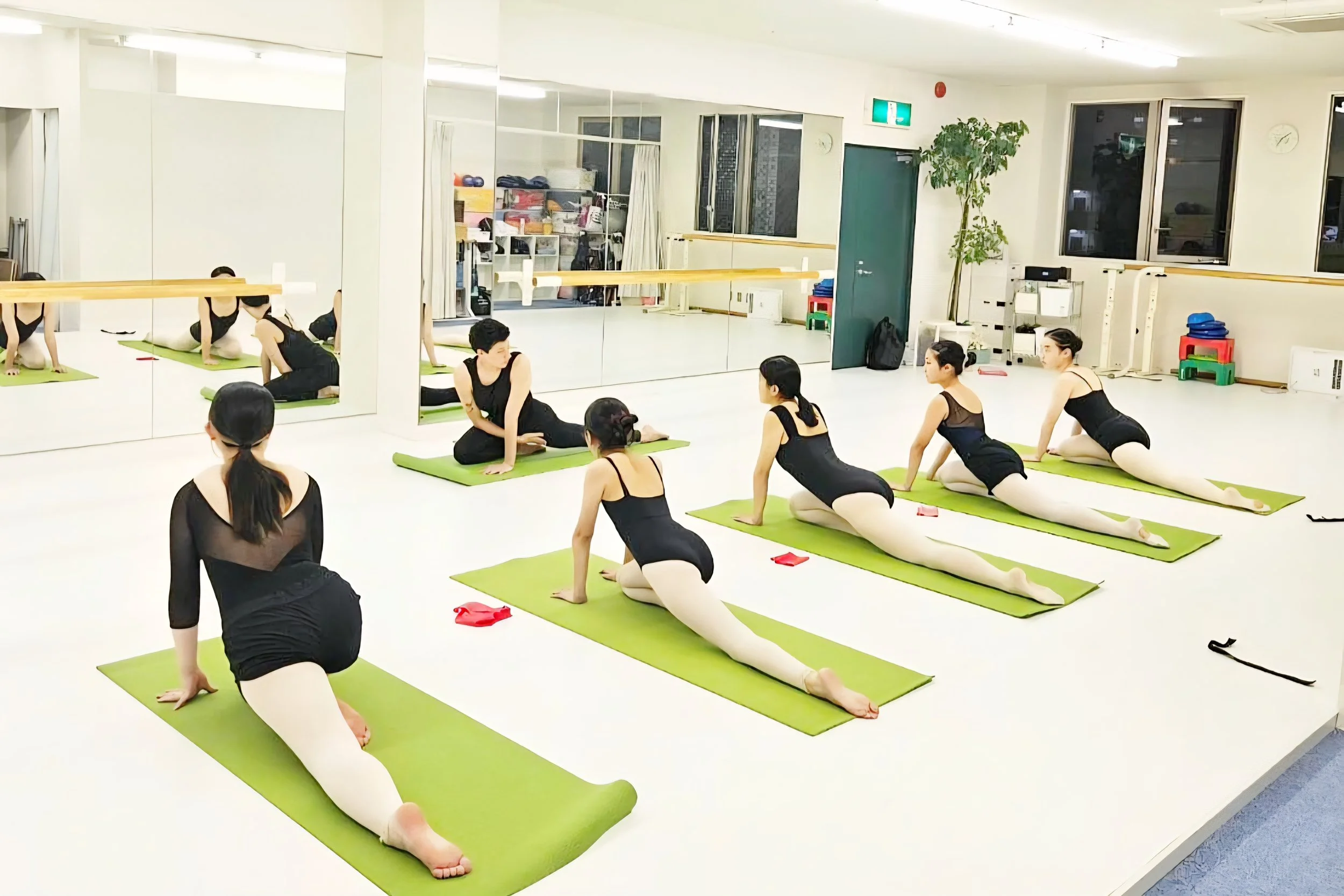 A group of young ballet dancers participating in a Body Tuning and conditioning class in a bright studio. They are practicing a stretch on green mats, wearing black top and white tights. The studio has mirrors, windows, and exercise equipment.