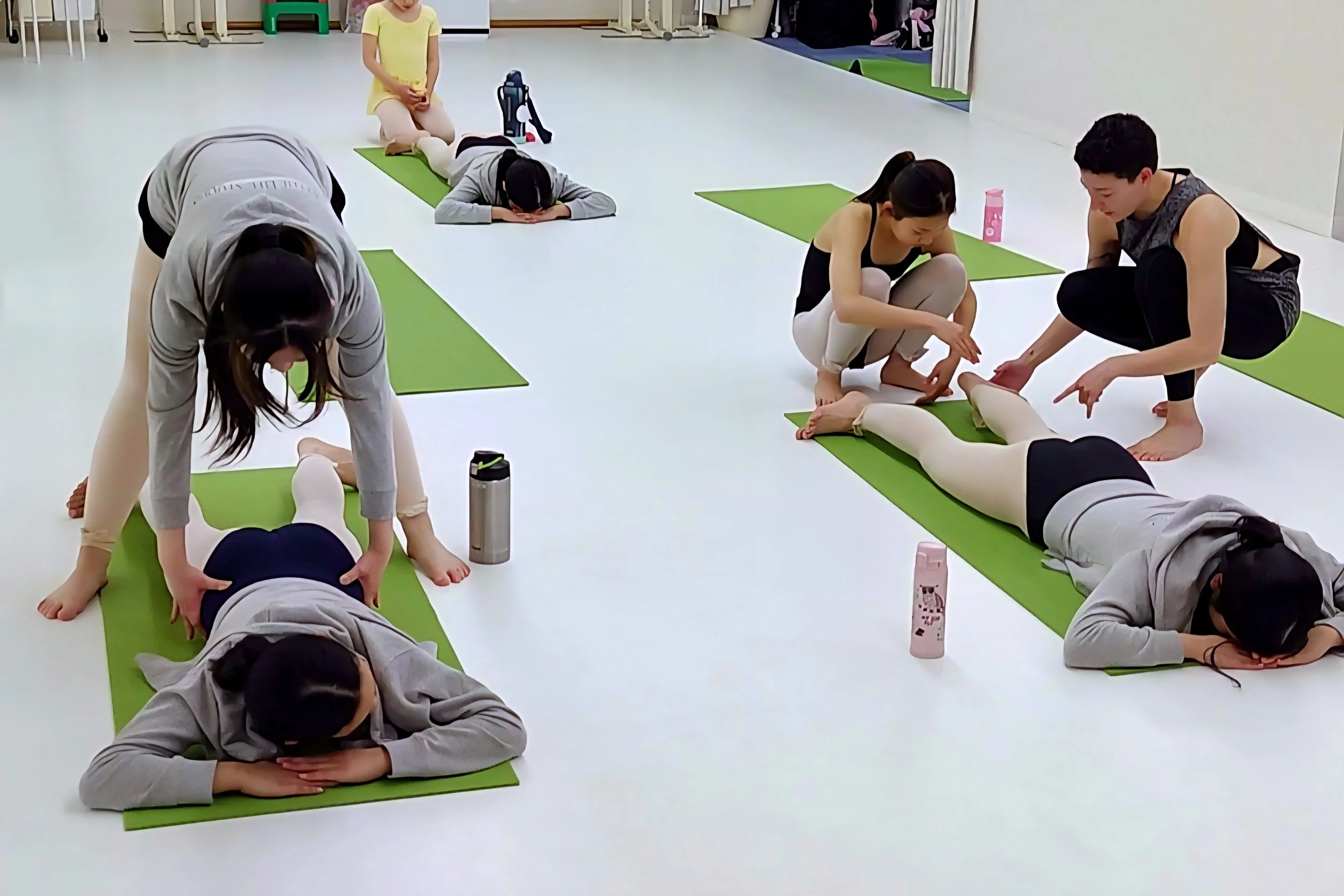 Young ballet students participating in a Body Tuning and conditioning class, lying on green mats, with instructors assisting them in a brightly lit indoor studio.