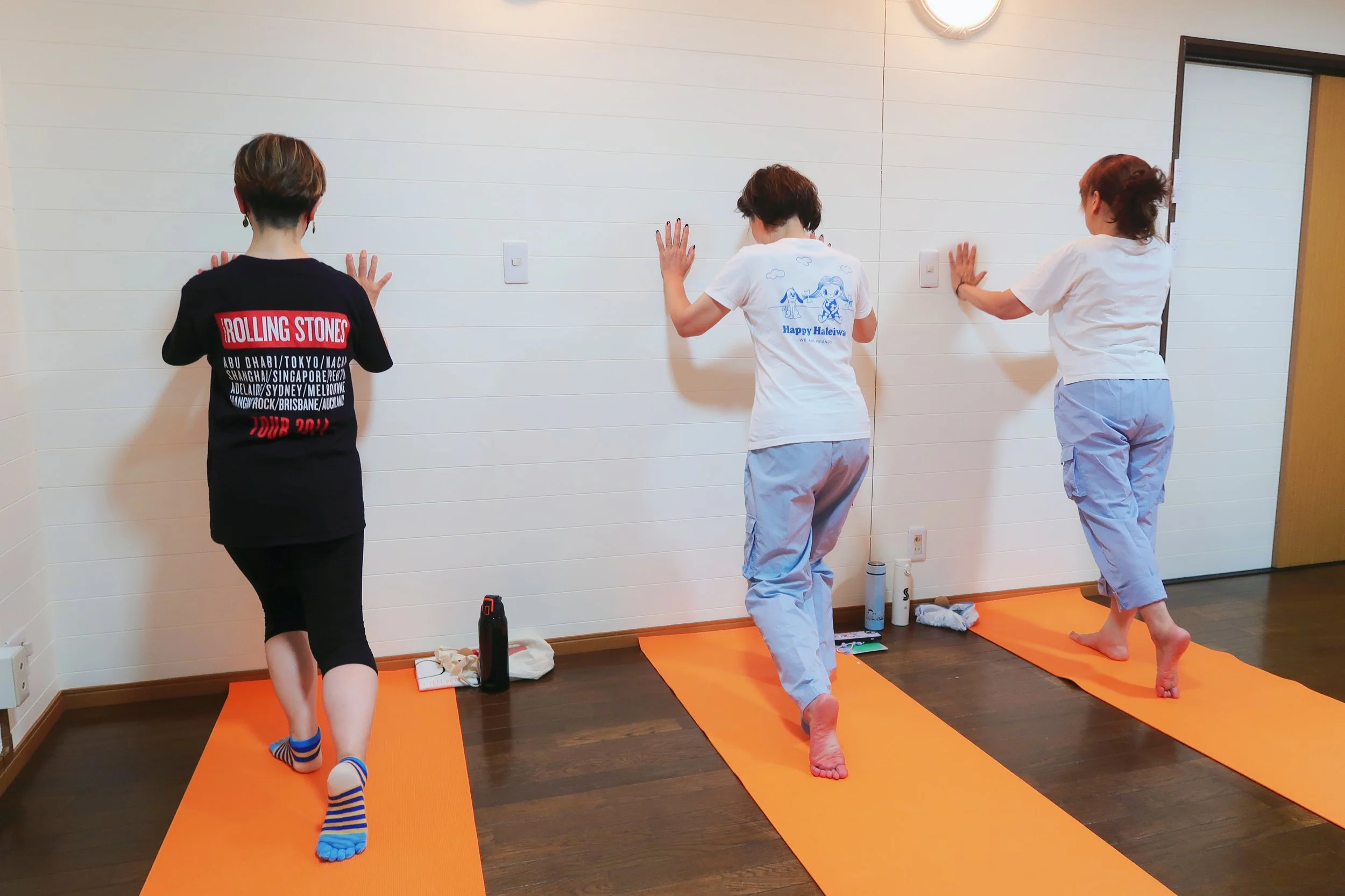 Three women practicing Body Tuning on orange mats in a studio with a white wall, wearing casual workout clothes, with water bottles and personal items beside them.