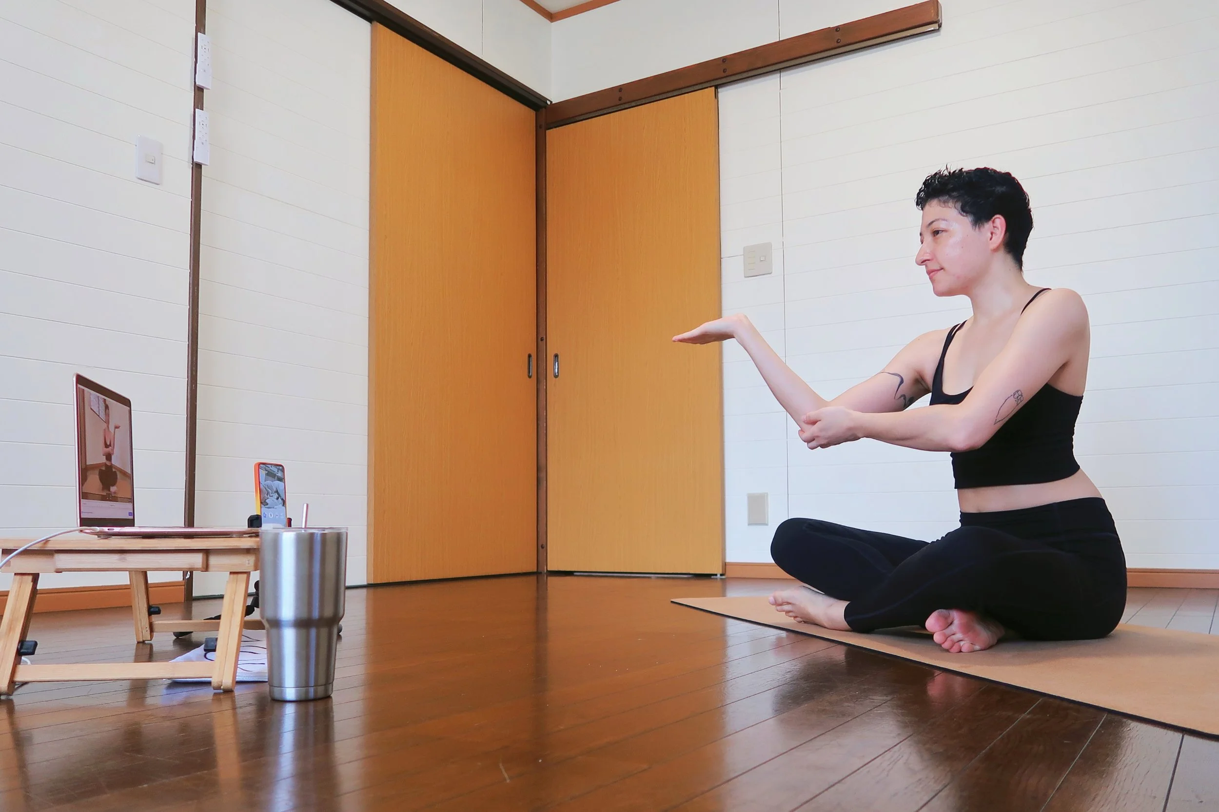 Body tuning instructor sits on her mat in a white studio while showing her online client how to loosen their shoulder blade. A silver tumbler sits next to the rose gold apple laptop