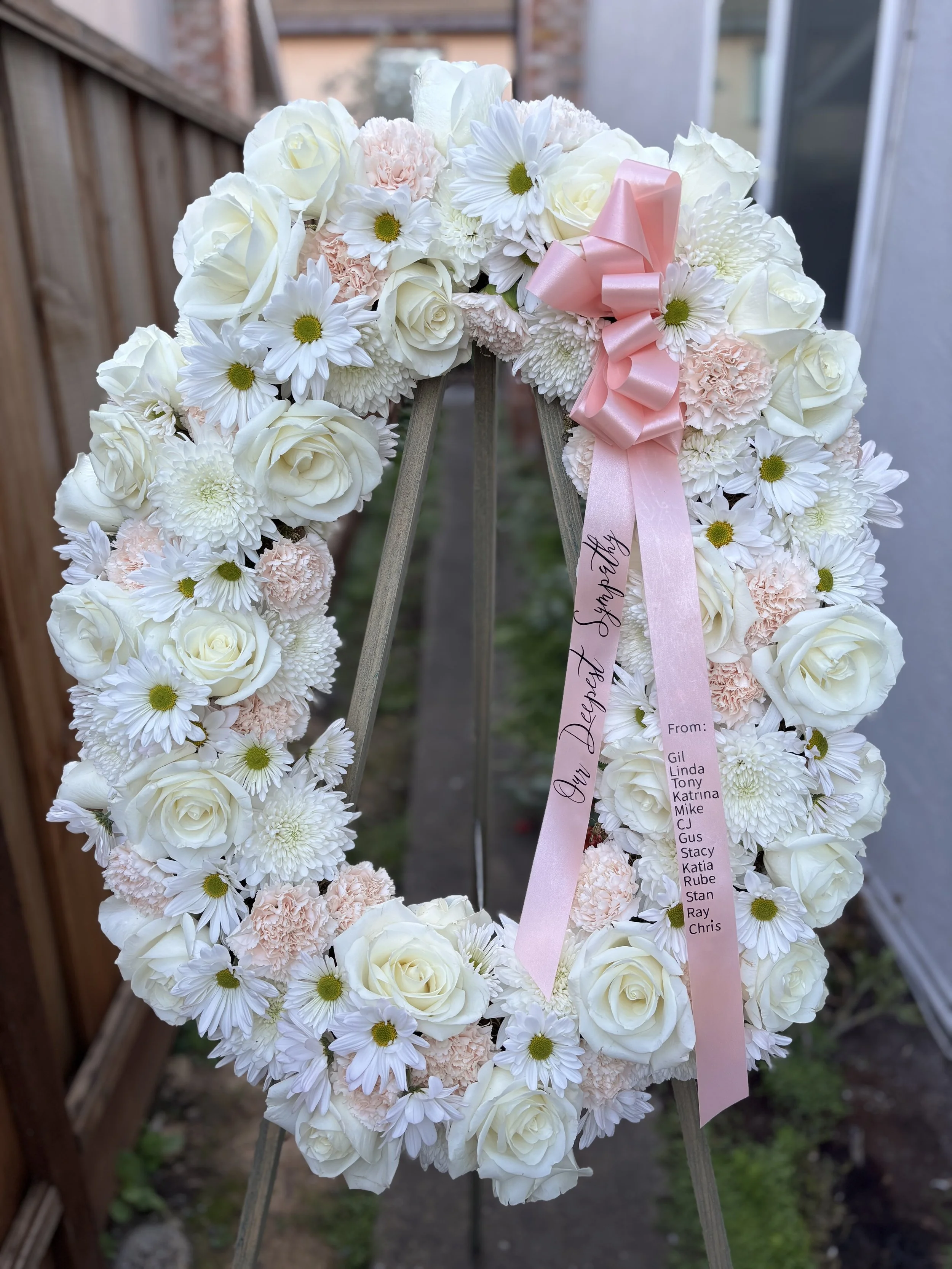A floral wreath made of white roses, daisies, chrysanthemums, and light pink carnations, decorated with a pink ribbon and a thank you note attached.