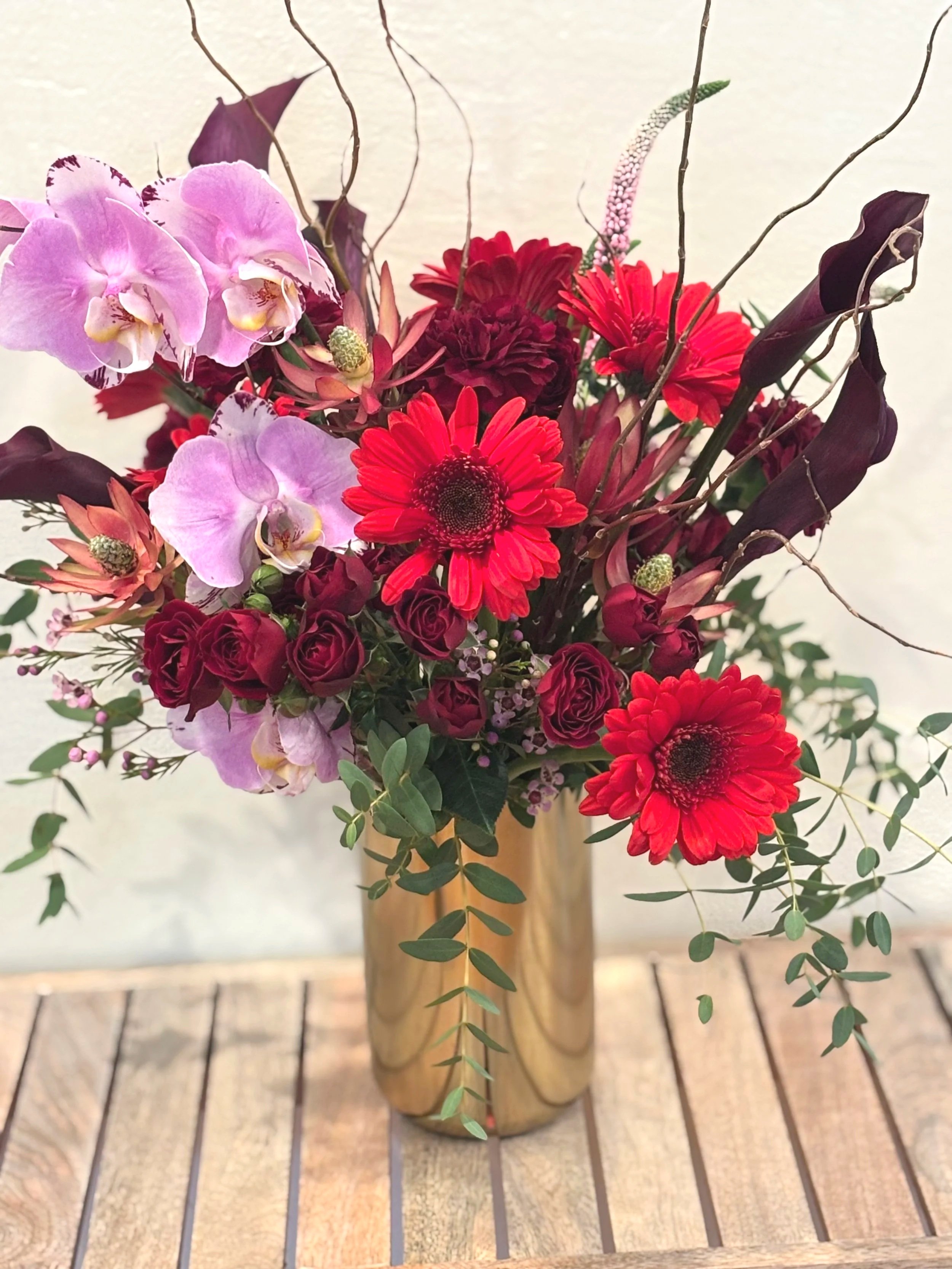 A floral arrangement in a shiny gold vase with pink orchids, red gerbera daisies, dark burgundy roses, and assorted greenery, placed on a wooden surface against a plain background.