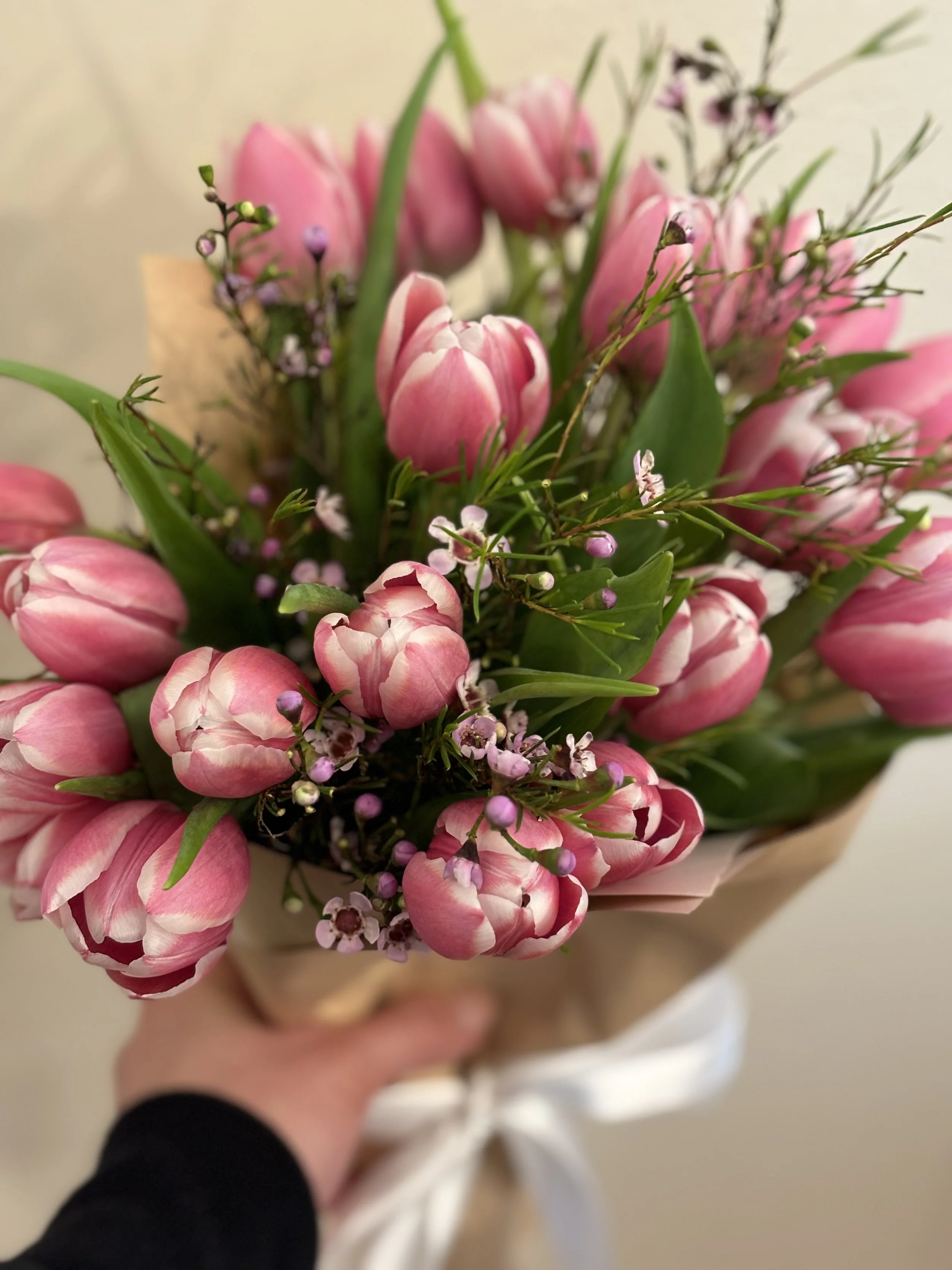 Hand holding a bouquet of pink and white tulips with small purple flowers wrapped in brown paper and tied with a white ribbon.