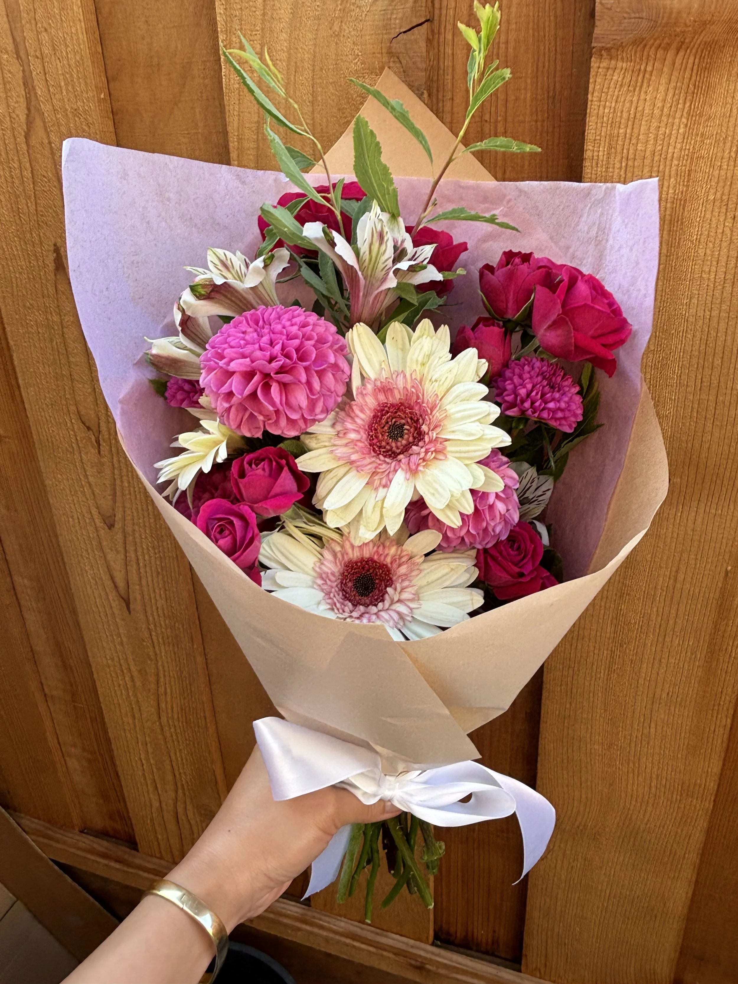 A hand holding a bouquet of pink and white flowers wrapped in beige paper with a white ribbon, set against a wooden background.