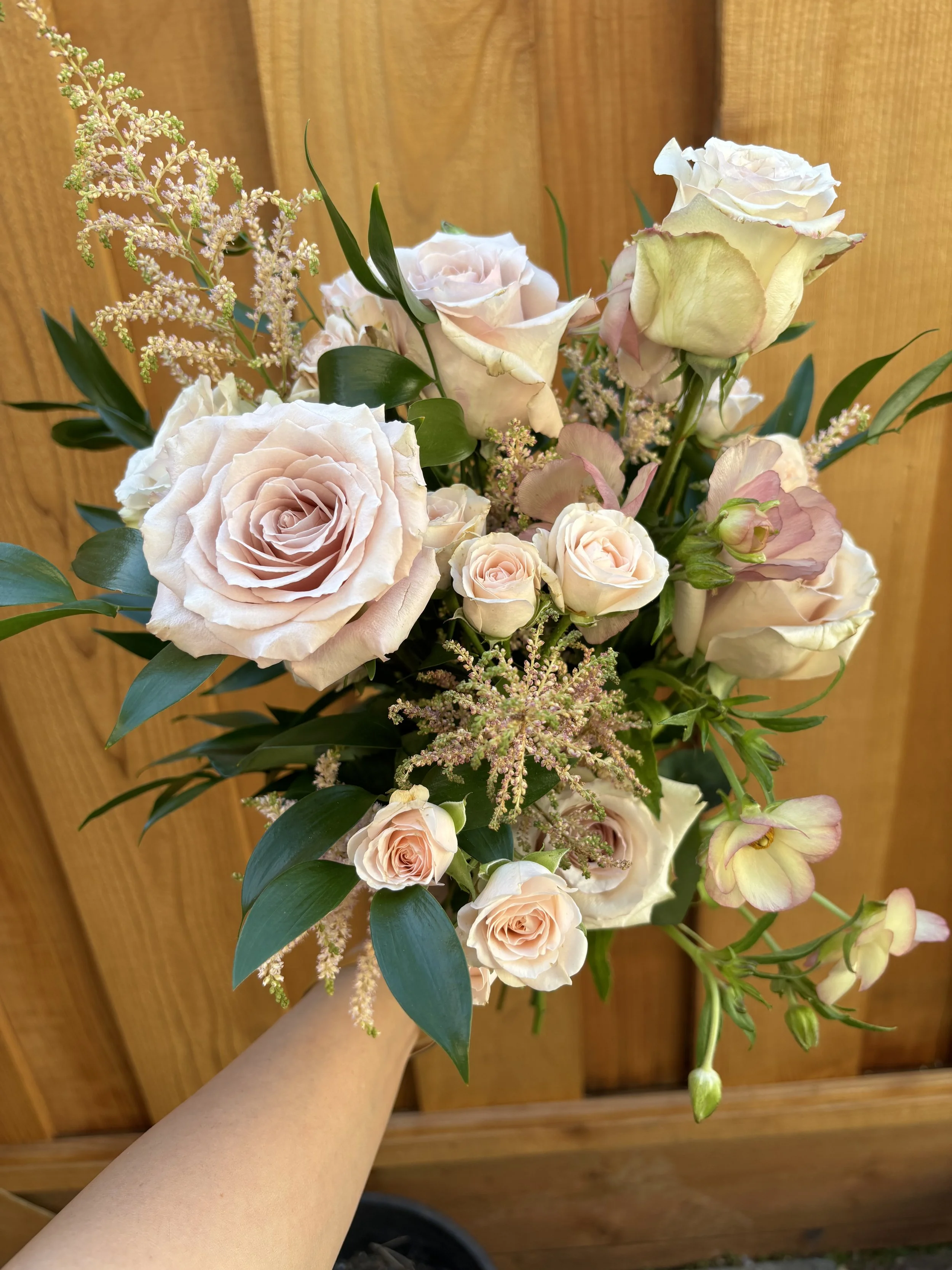 A hand holding a bouquet of light pink and white roses, with some greenery and small filler flowers, against a wooden background.