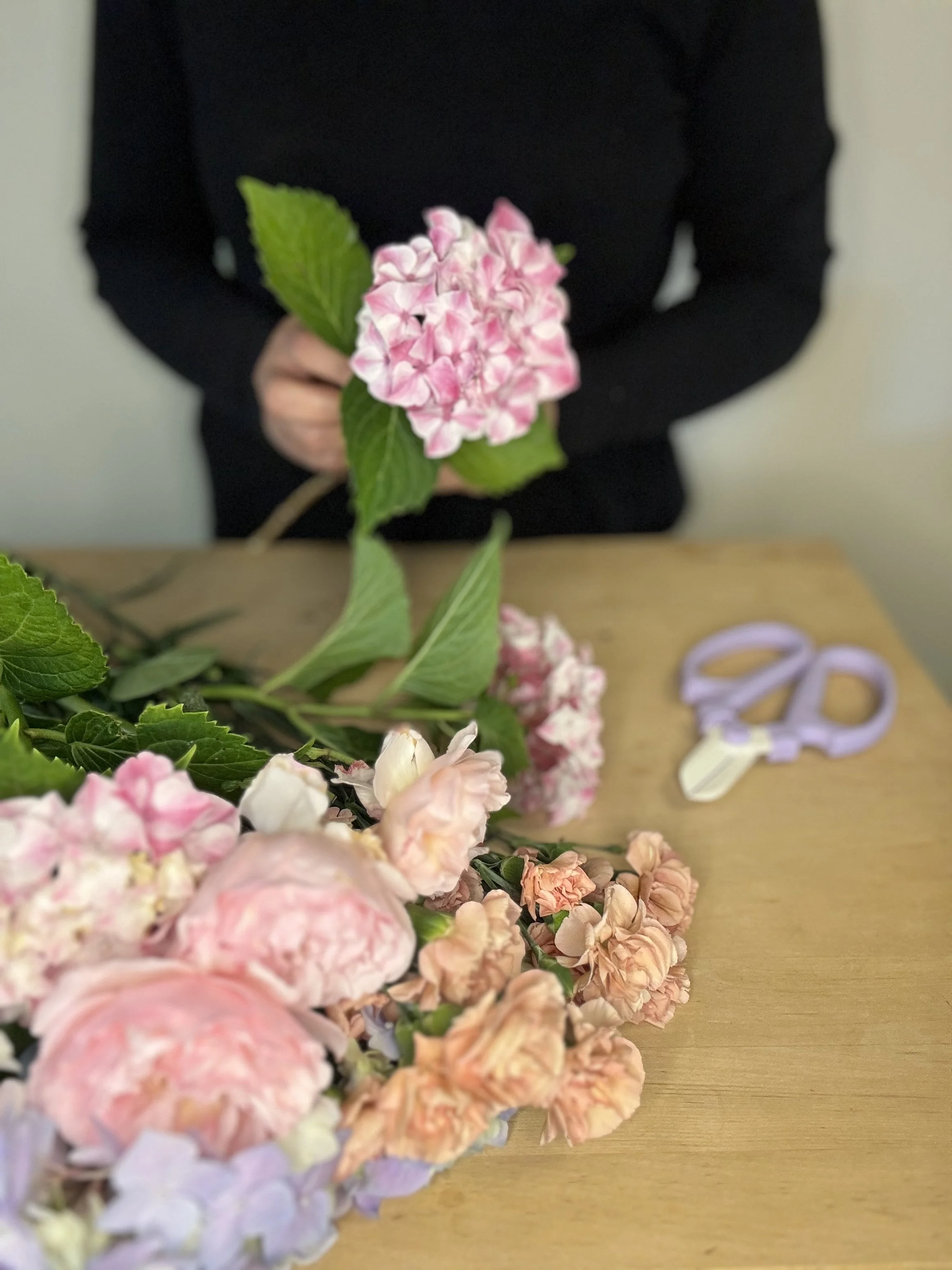 Person arranging pink and light purple hydrangea flowers on a wooden table, with scissors nearby.