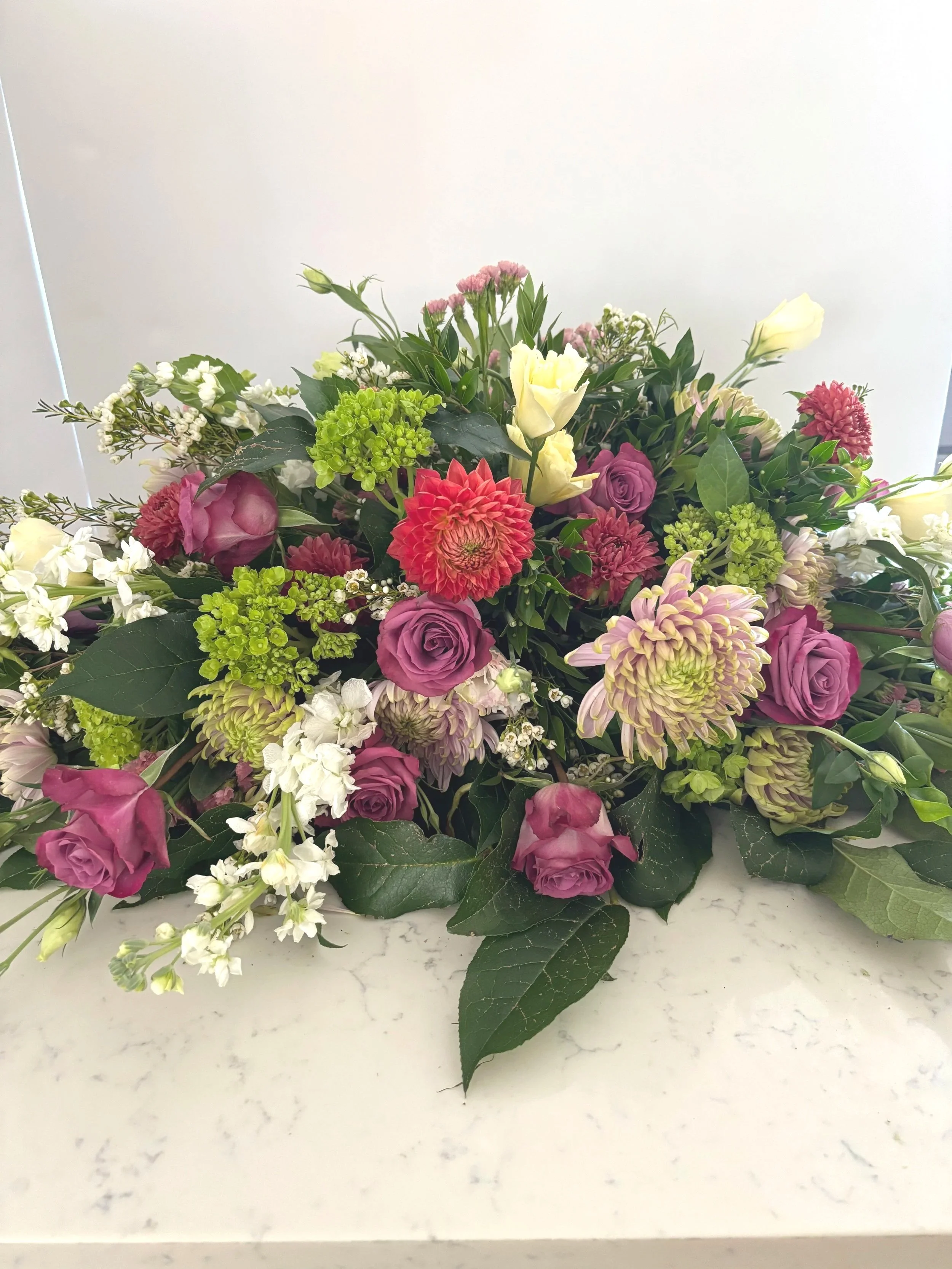 A colorful bouquet of various flowers including roses, dahlias, and lisianthus, accented with greenery, resting on a white surface.