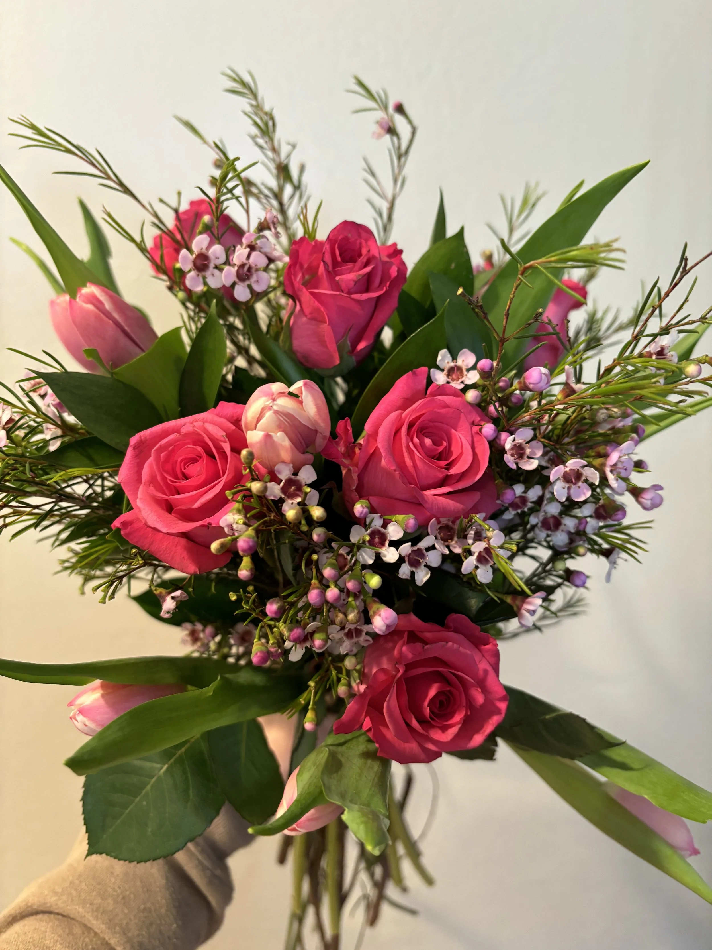 A hand holding a bouquet of pink roses, small white and pink flowers, and green leaves.